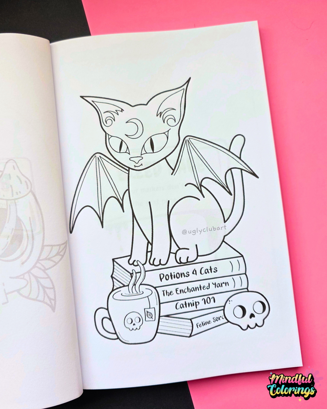 An open coloring book showing a black-and-white line art page of a bat-winged cat sitting on books labeled 'Potions & Cats', placed on a pink background with a black section visible behind it.