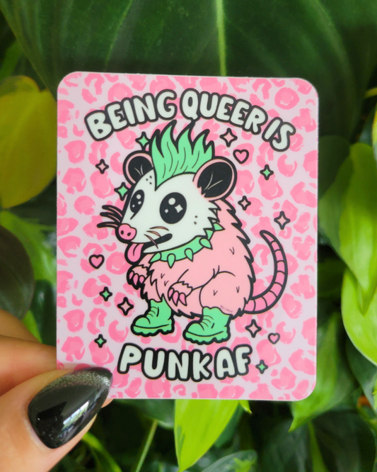 A pink possum character with it's tongue out, a green mohawk and wearing a green spikey necklace against a pink cheetah print background with the words "Being Queer is Punk AF"