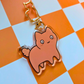 Acrylic keychain featuring a chubby orange cat  looking concerned with wide eyes attached to a gold cat-shaped clasp on an orange checkered background.