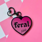 Pink heart-shaped acrylic keychain featuring the word "feral" and "xoxo" with a black heart clasp on a pink checkered background.