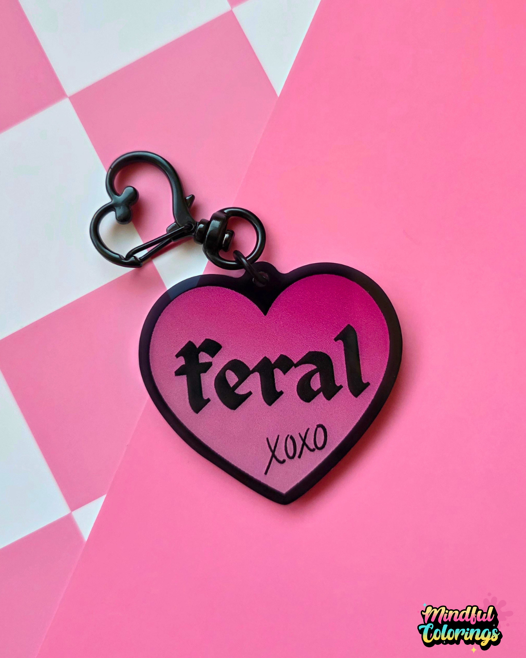 Pink heart-shaped acrylic keychain featuring the word "feral" and "xoxo" with a black heart clasp on a pink checkered background.