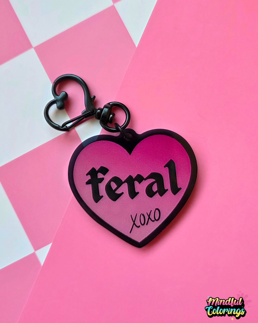 Pink heart-shaped acrylic keychain featuring the word "feral" and "xoxo" with a black heart clasp on a pink checkered background.