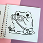 Mini frog-themed colouring book page featuring a curious frog looking through binoculars while carrying a small satchel.