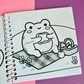Mini frog-themed colouring book page featuring a happy frog enjoying a picnic while eating a sandwich on a checkered blanket surrounded by flowers and a picnic basket.