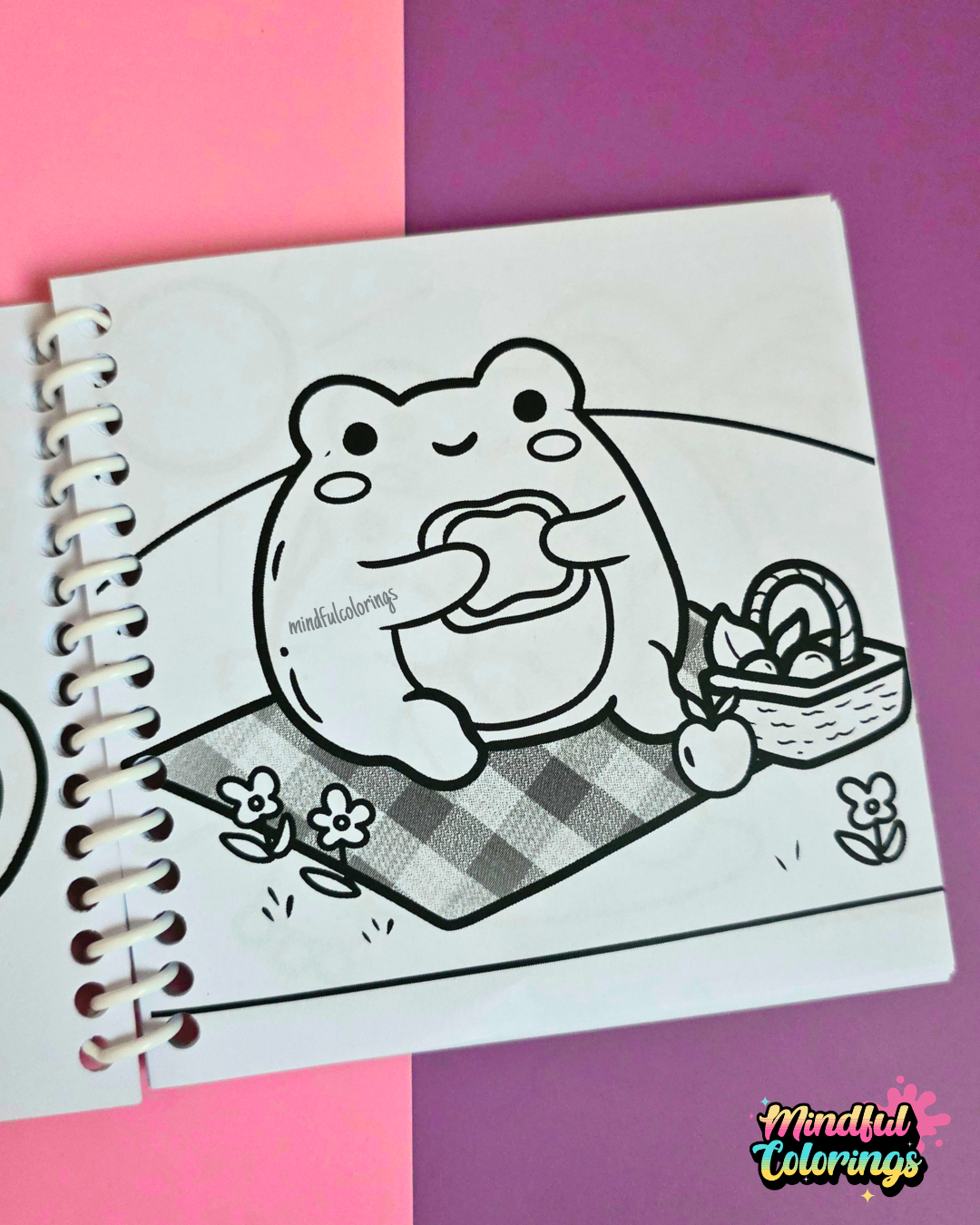 Mini frog-themed colouring book page featuring a happy frog enjoying a picnic while eating a sandwich on a checkered blanket surrounded by flowers and a picnic basket.