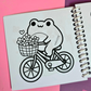 Mini frog-themed colouring book page featuring a happy frog riding a bicycle with a basket full of flowers.