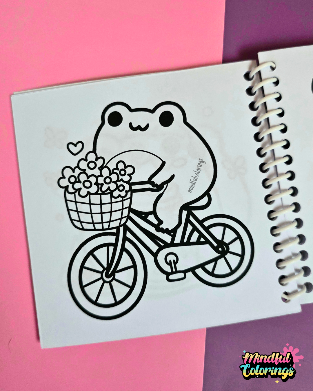 Mini frog-themed colouring book page featuring a happy frog riding a bicycle with a basket full of flowers.