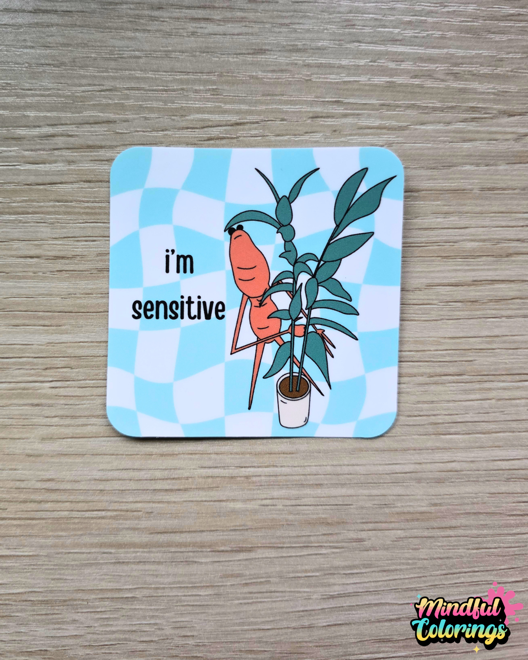 Sticker with the viral Marcus the Worm character hiding behind a green houseplant with the text "I'm Sensitive" with a blue checkered background