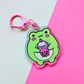 Acrylic keychain featuring a cute green frog holding a pink bubble tea drink on a pink and mint background.