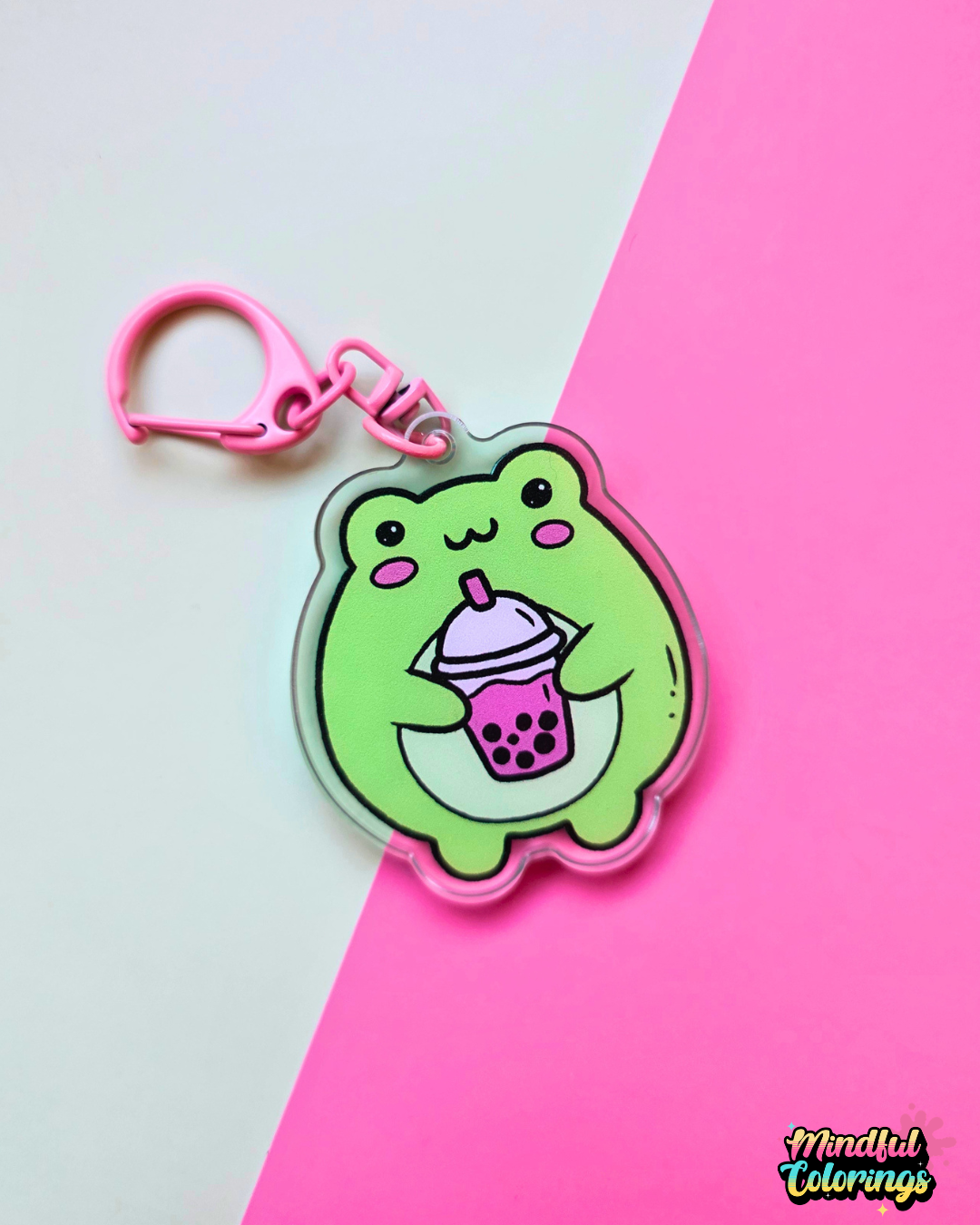 Acrylic keychain featuring a cute green frog holding a pink bubble tea drink on a pink and mint background.