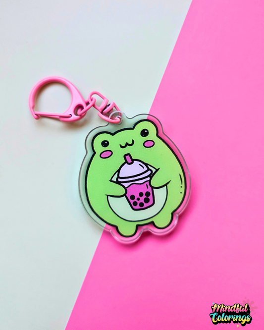 Acrylic keychain featuring a cute green frog holding a pink bubble tea drink on a pink and mint background.
