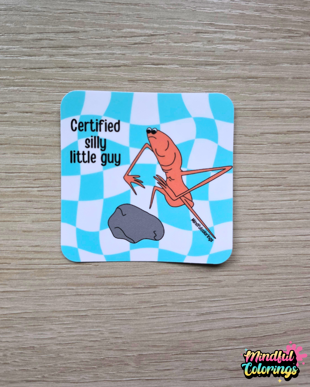 Sticker with the viral Marcus the Worm character crouched next to a rock with the text "Certified Silly Little Guy" with a blue checkered background