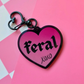 Pink heart-shaped acrylic keychain featuring the word "feral" and "xoxo" with a black heart clasp on a pink checkered background.