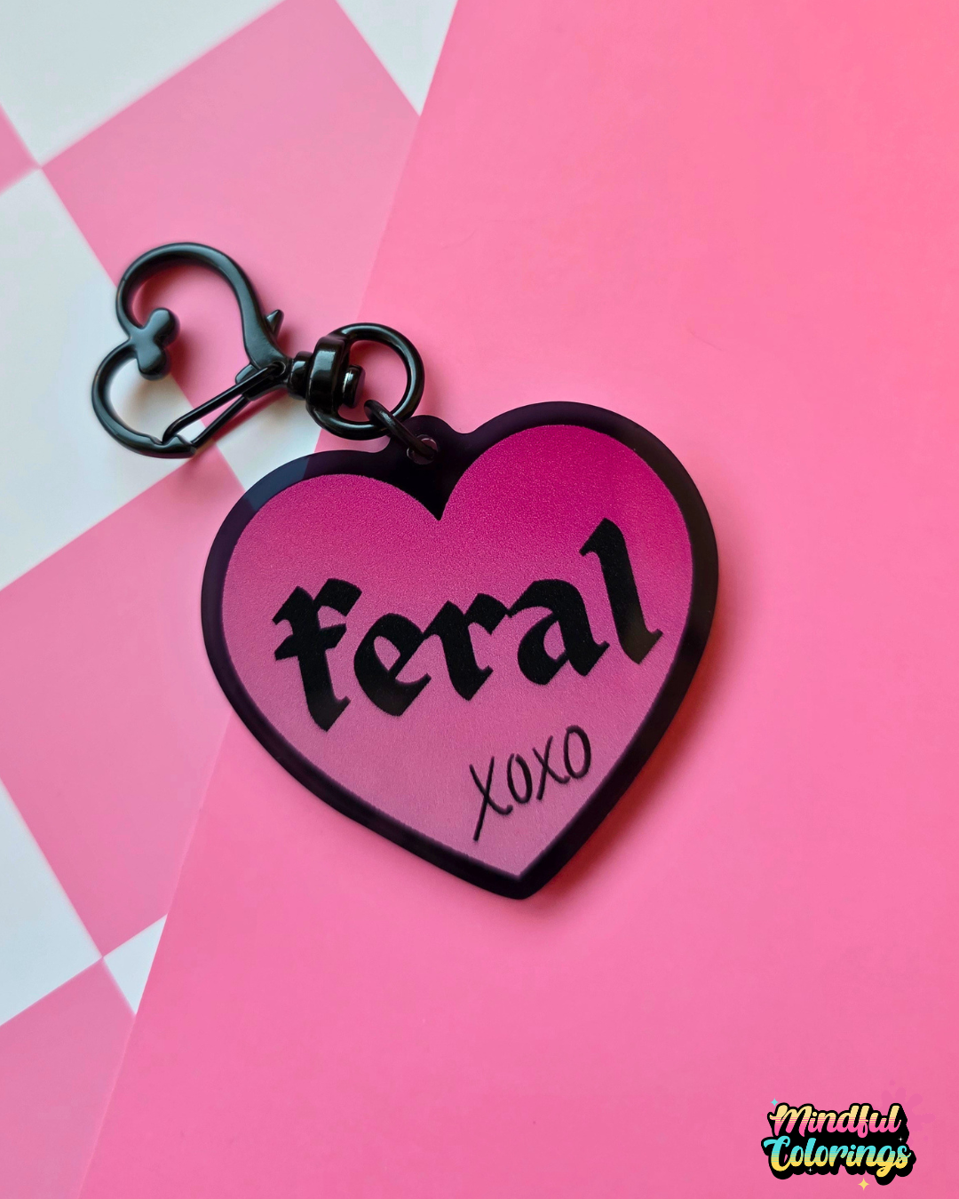 Pink heart-shaped acrylic keychain featuring the word "feral" and "xoxo" with a black heart clasp on a pink checkered background.