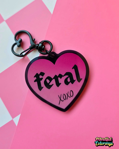 Pink heart-shaped acrylic keychain featuring the word "feral" and "xoxo" with a black heart clasp on a pink checkered background.