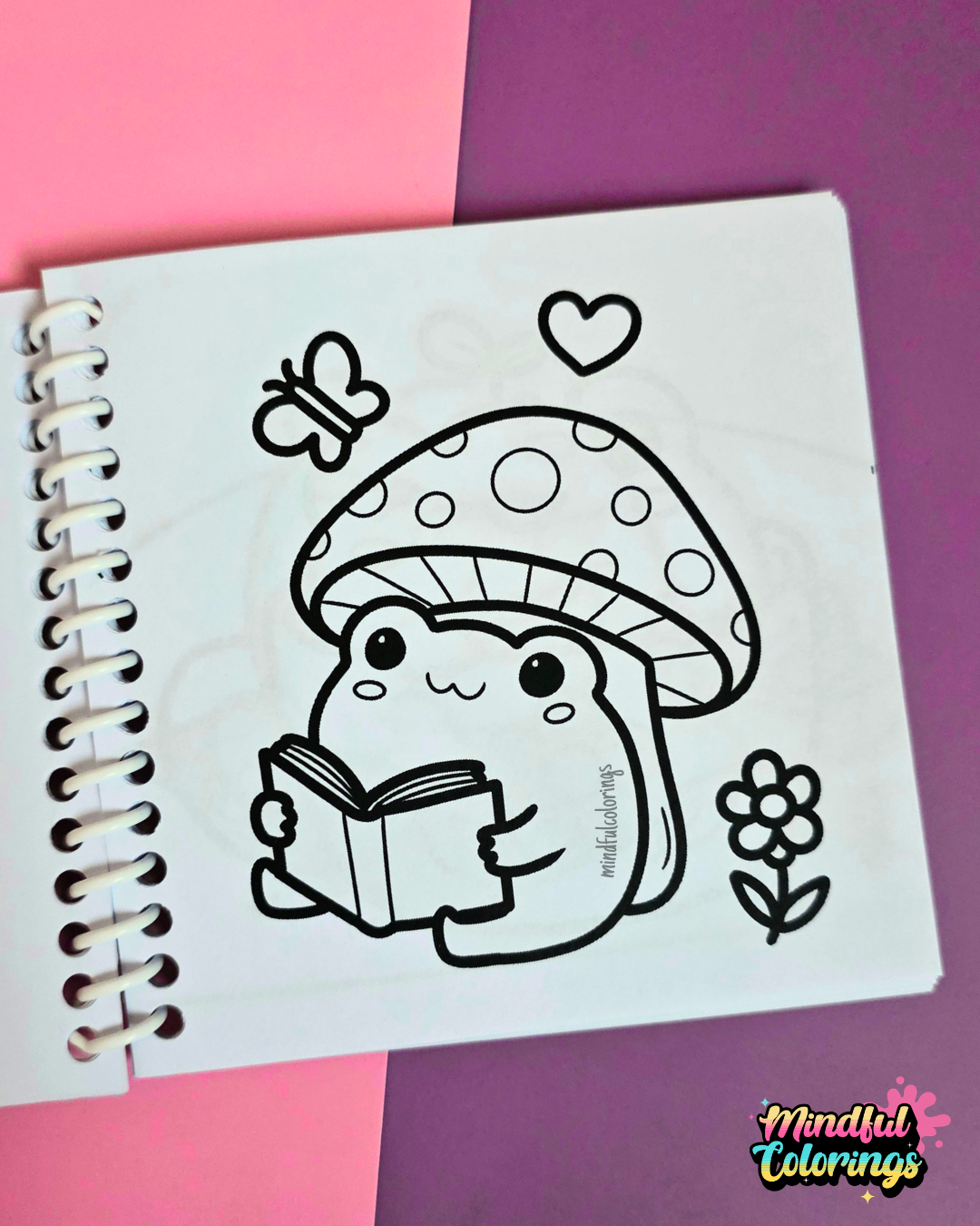Mini frog-themed colouring book page featuring a cute frog reading a book while sitting beneath a spotted mushroom, surrounded by a butterfly, flower, and heart.