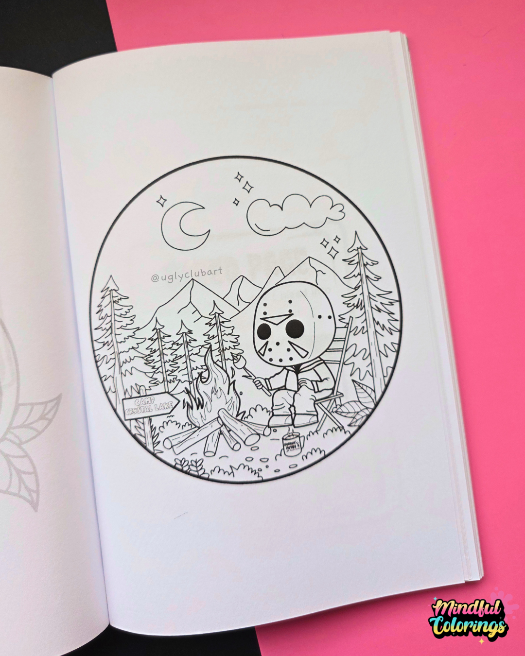 An open coloring book showing a black-and-white circular campsite scene with a masked figure, trees, mountains, and a campfire. The page is placed on a bright pink background, with a small 'Mental Colorings' logo in the bottom right corner.