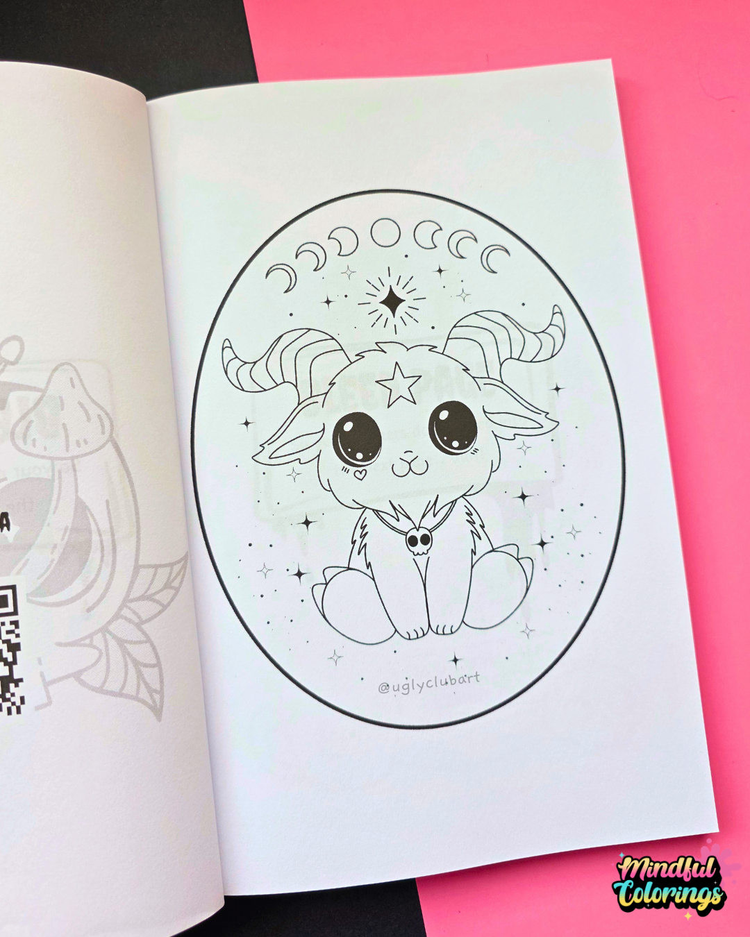 An open coloring book or art booklet showing a black-and-white line drawing of a cute baphomet inside an oval frame, placed on a bright pink background with part of another page visible on the left.