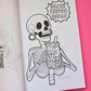An open coloring book or art print showing a black-and-white line drawing of a skeleton holding a drink, with the text 'DEATH BEFORE DECAF' on the page. The book is photographed on a bright pink background with a small 'Mindful Colorings' logo in the lower right corner.