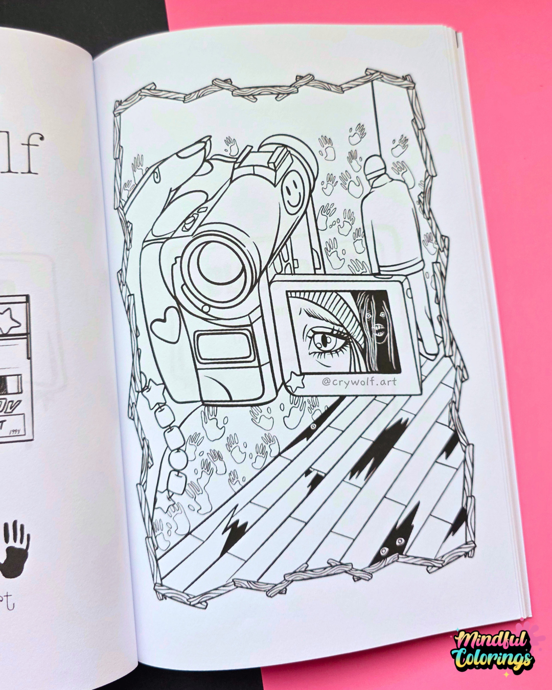 An open coloring book or activity booklet showing a black-and-white illustrated page with a camera, a phone screen with Blair Witch Movies background, placed on a bright pink background.