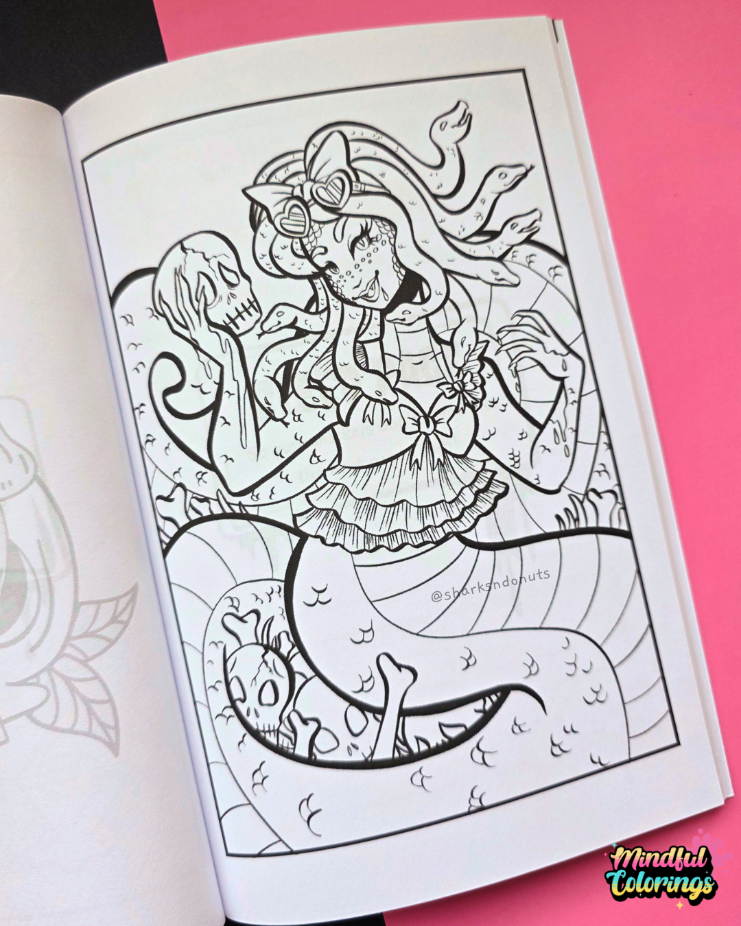 An open coloring book showing a black and white line art illustration of a Medusa with a skull on her head, placed on a bright pink background.