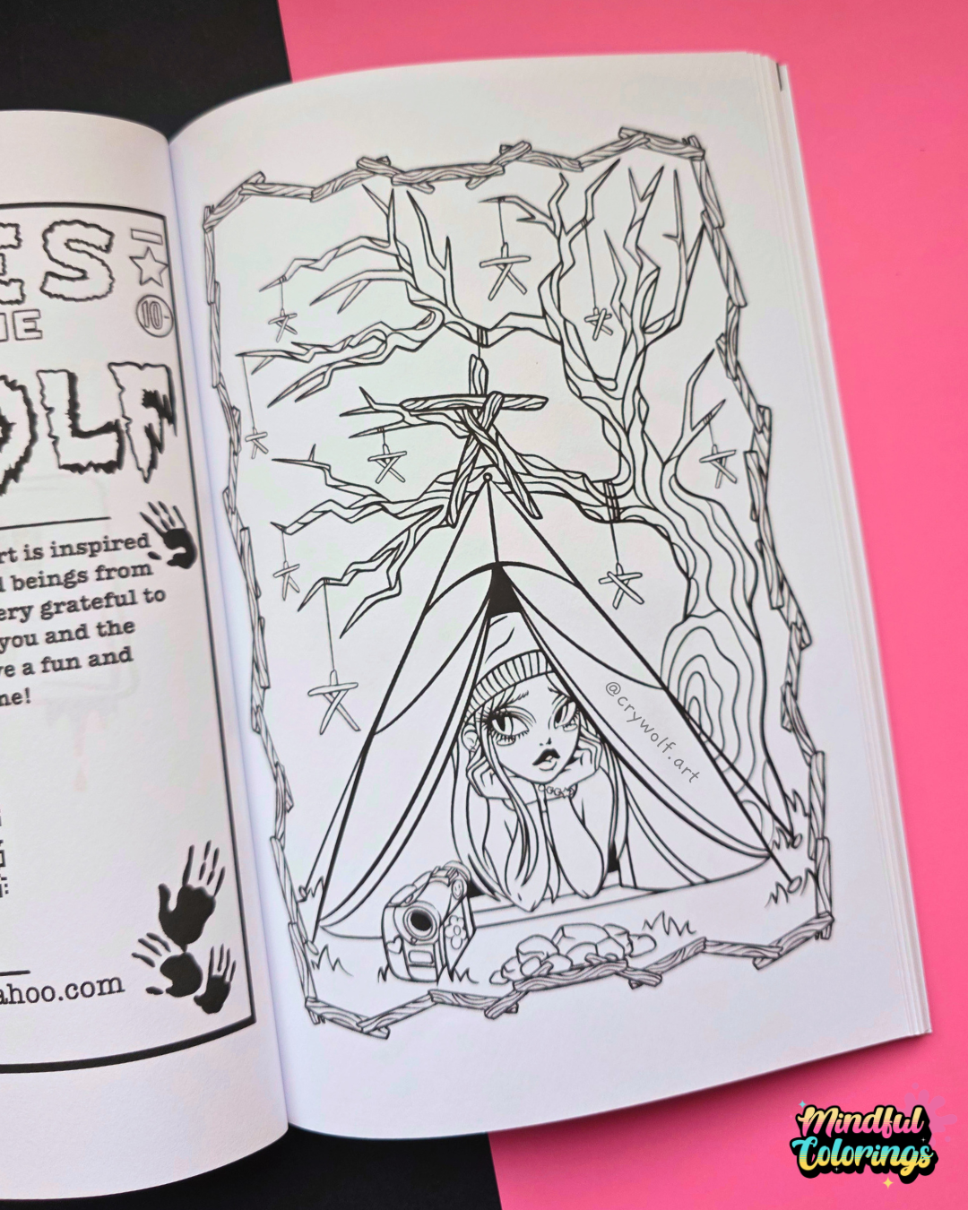 An open coloring book showing a black and white line art illustration of a girl sitting inside a tent under a tree with Blair Witch Movie totems. The book is placed on a bright pink background with a black section visible on the left.
