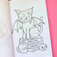 An open coloring book showing a black-and-white line art page of a bat-winged cat sitting on books labeled 'Potions & Cats', placed on a pink background with a black section visible behind it.