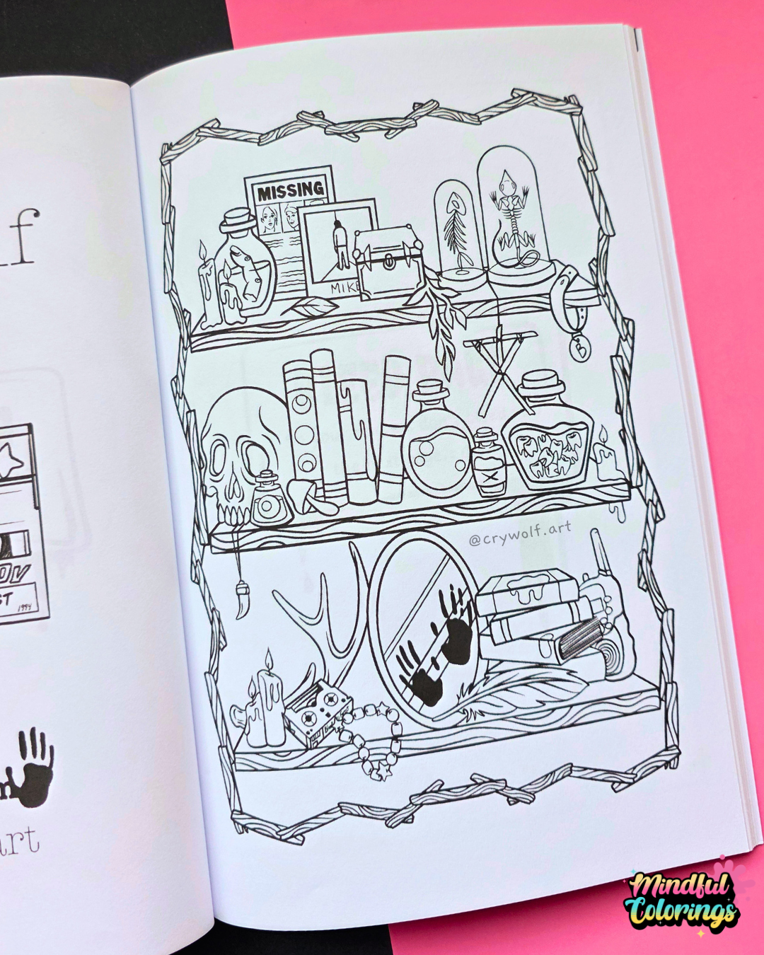 An open coloring book or art booklet showing a black-and-white illustrated page with shelves of occult and gothic objects like a skull, cauldron, books, and mirrors, placed against a bright pink background.