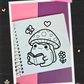 Adorable frog reading a book beneath a spotted mushroom coloring page with butterfly and flower details from the Frogcore Adventures mini coloring book.